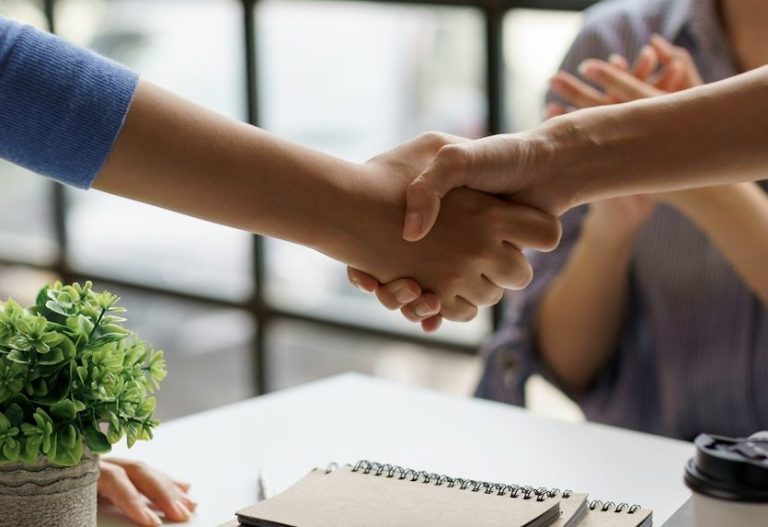 Two hands shaking over a pile of notebooks while a third person looks on, smiling